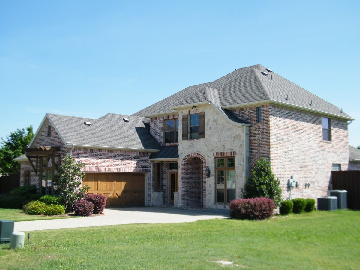Large two-story brick and stone home featuring rustic wooden garage doors, an arched entryway, and a green front lawn, representing residential properties inspected by Northmark Property Inspections in Suamico, WI.